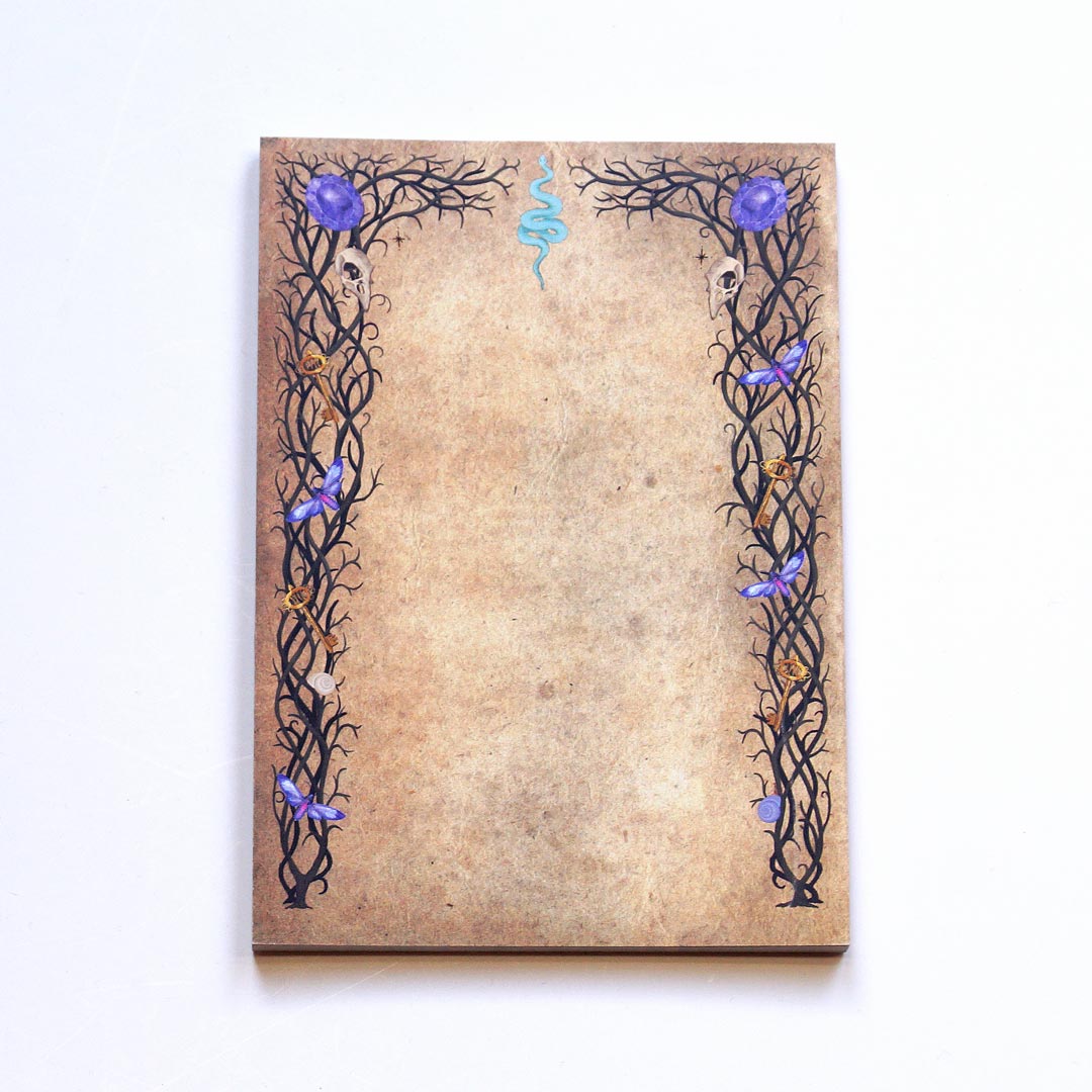 A witchy notepad with 50 sheets of double-sided printed paper, featuring an aged parchment-like background. The intricate black vine-like borders are adorned with small skulls, purple crystals, golden keys, and delicate blue butterflies. A blue serpent symbol decorates the top center. The notepad is photographed on a white surface, showcasing its mystical and gothic-inspired design.