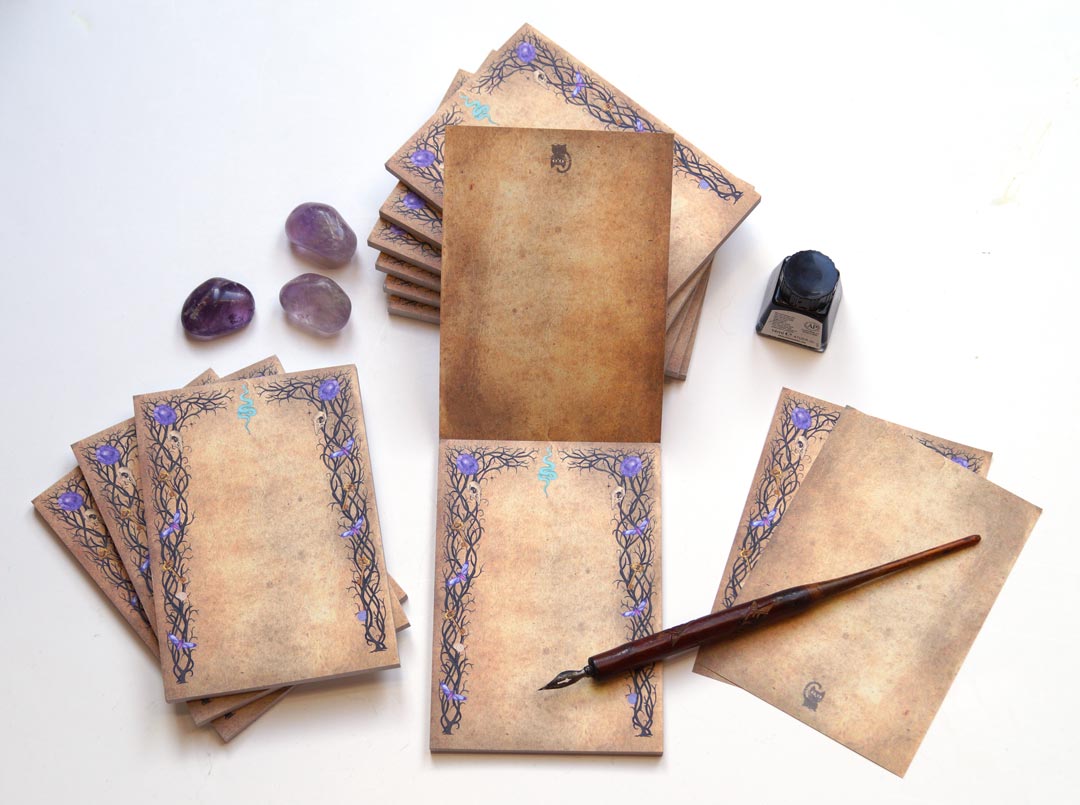 A witchy, gothic-inspired notepad with 50 sheets of double-sided printed paper, featuring an aged parchment-like background. The intricate black vine-like borders are adorned with small skulls, purple crystals, golden pentagrams, and a blue serpent symbol at the top center. The notepad is displayed with multiple stacks, some open to reveal the double-sided print, alongside amethyst crystals, a vintage dip pen, and a bottle of black ink on a white surface, evoking a mystical and enchanting aesthetic.