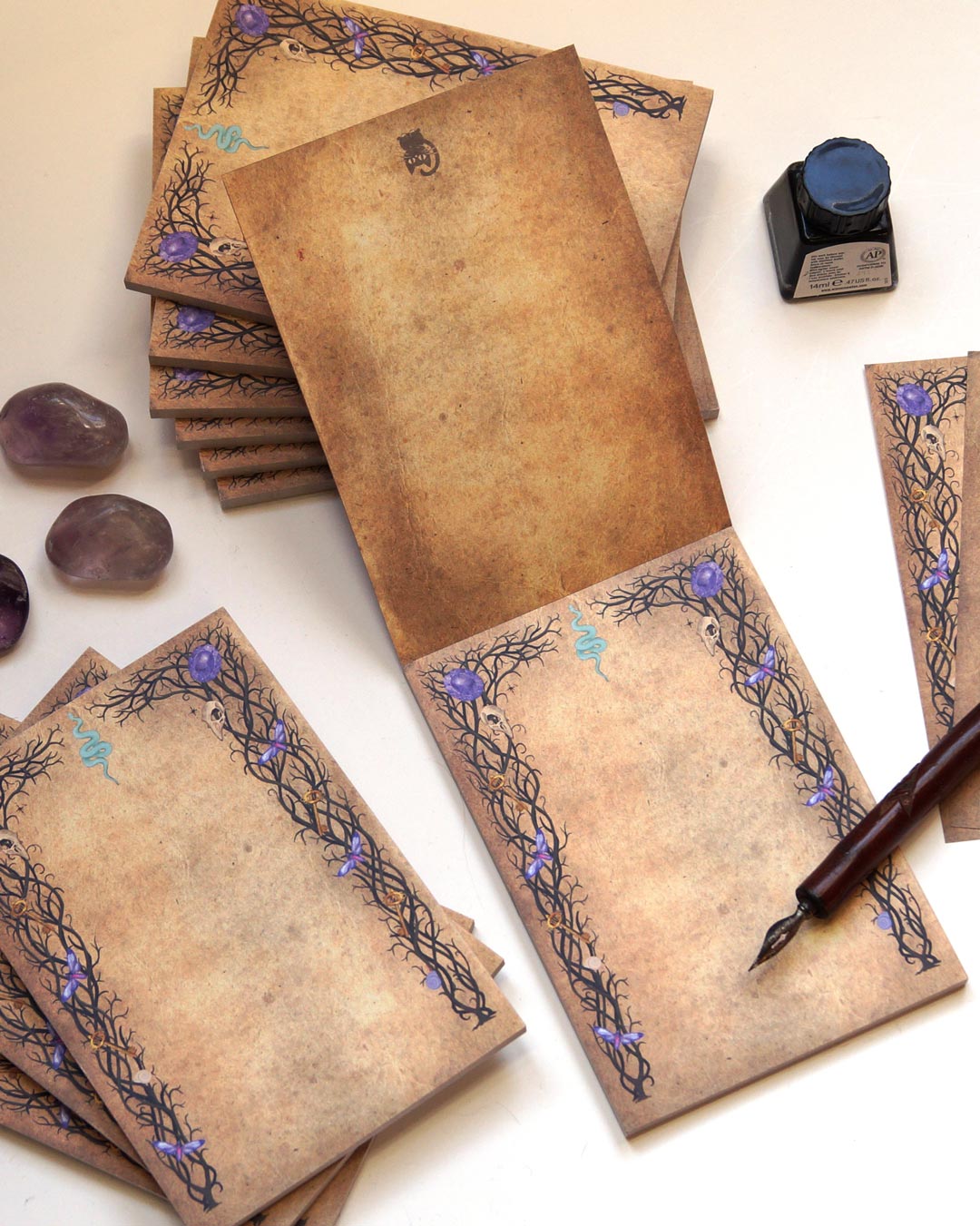 A witchy, gothic-inspired notepad with 50 sheets of double-sided printed paper, featuring an aged parchment-like background. The intricate black vine-like borders are adorned with small skulls, purple crystals, golden pentagrams, and a blue serpent symbol at the top center. The notepad is displayed with multiple stacks, some open to reveal the double-sided print, alongside amethyst crystals, a vintage dip pen, and a bottle of black ink on a white surface, evoking a mystical and enchanting aesthetic.