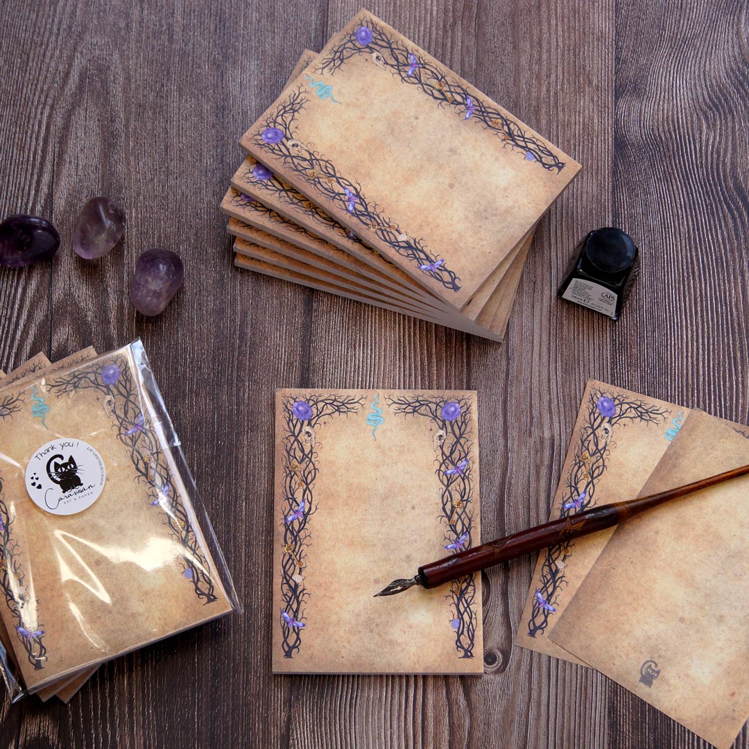 A witchy, gothic-inspired notepad with 50 sheets of double-sided printed paper, featuring an aged parchment-like background. The intricate black vine-like borders are decorated with small skulls, purple crystals, golden keys, and a blue serpent symbol at the top center. The notepad is displayed on a rustic wooden surface, accompanied by a vintage dip pen, a bottle of black ink, and amethyst crystals. Stacks of notepads are neatly arranged, with one wrapped in protective plastic, creating a mystical statione