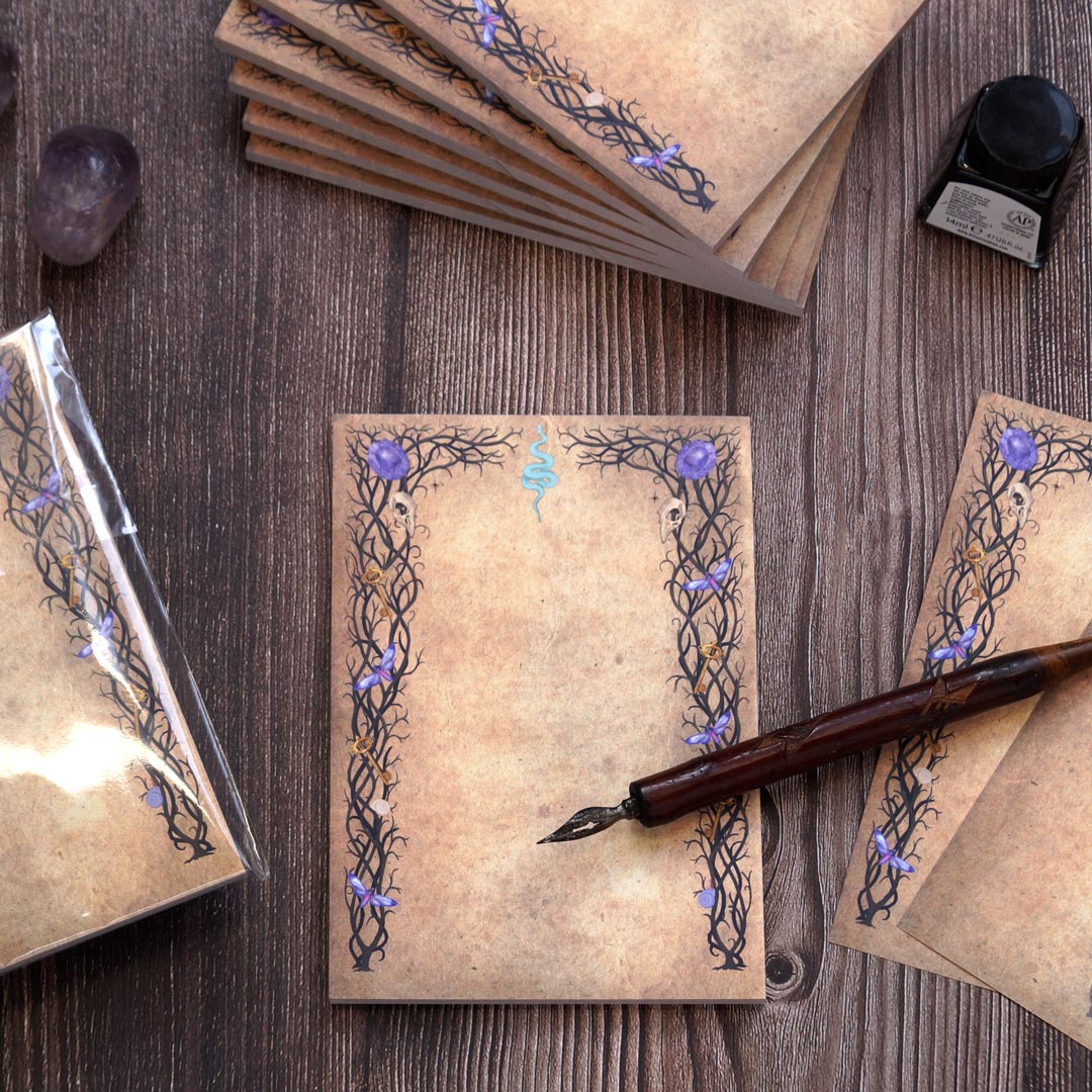 A witchy, gothic-inspired notepad with 50 sheets of double-sided printed paper, featuring an aged parchment-like background. The intricate black vine-like borders are decorated with small skulls, purple crystals, golden keys, and a blue serpent symbol at the top center. The notepad is displayed on a rustic wooden surface, accompanied by a vintage dip pen, a bottle of black ink, and amethyst crystals. Stacks of notepads are neatly arranged, with one wrapped in protective plastic, creating a mystical statione