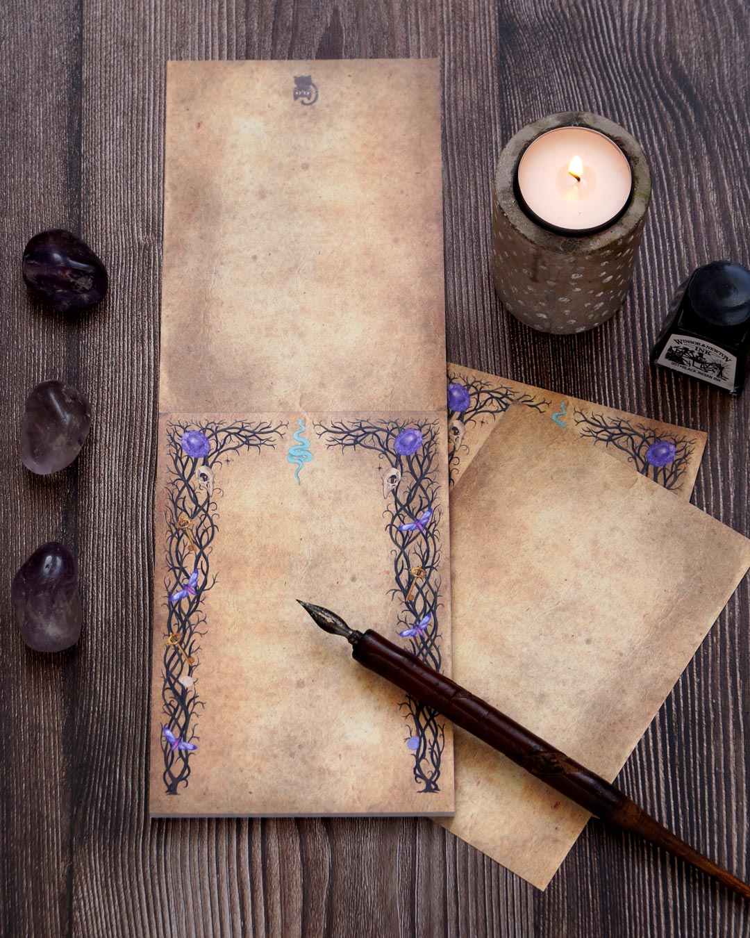 A witchy, gothic-inspired notepad with 50 sheets of double-sided printed paper, featuring an aged parchment-like background. The intricate black vine-like borders are adorned with small skulls, purple crystals, golden pentagrams, and a blue serpent symbol at the top center. The notepad is displayed open on a rustic wooden surface, revealing the back side of the paper with a small black cat symbol. A vintage dip pen rests on top, alongside amethyst crystals, a lit candle in a stone holder, and a bottle of bl
