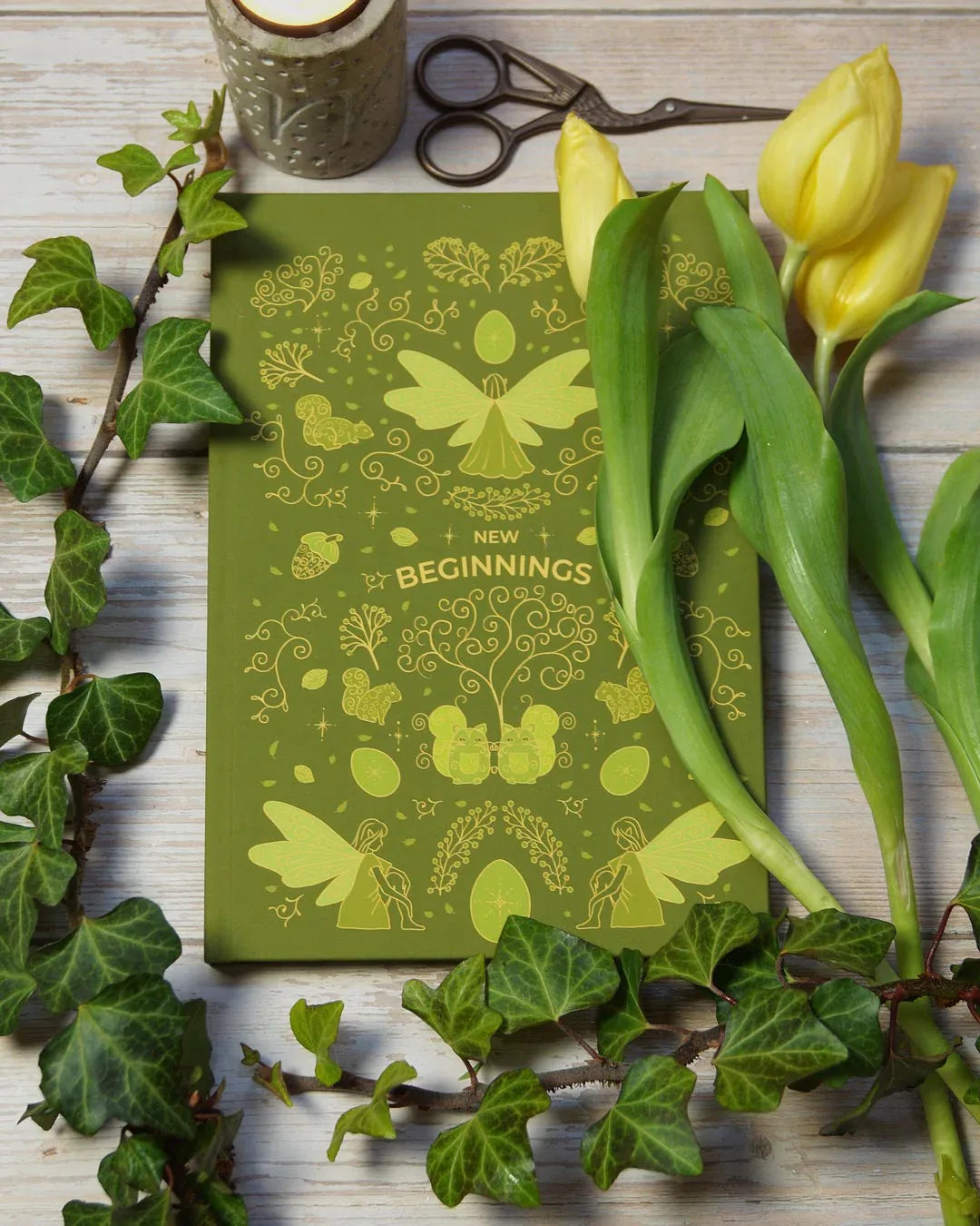 Green fairy-inspired hardcover journal with stylized ornamental illustrations photographed on a white wooden table beside fresh yellow tulips in soft natural daylight.