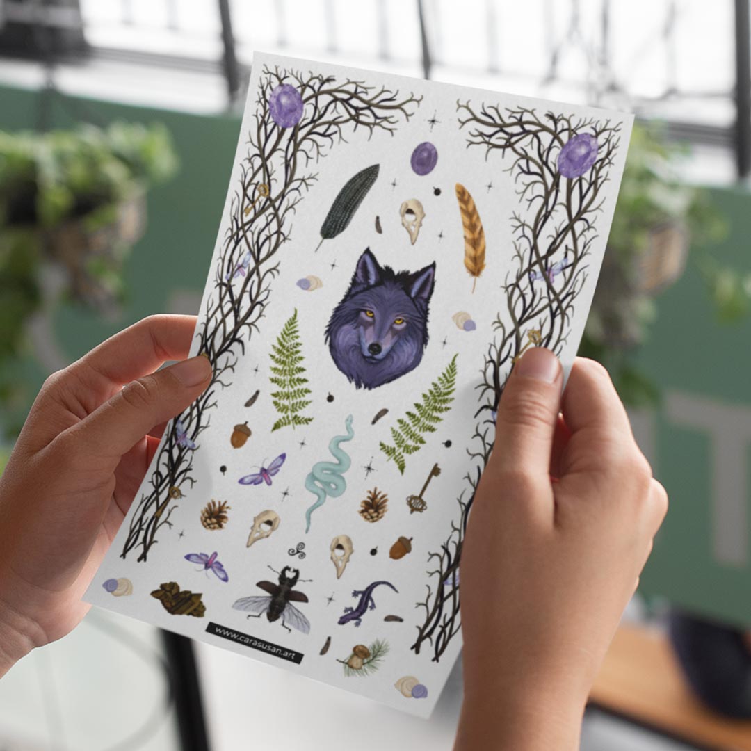 A person holds a sheet of stickers designed for bullet journaling and decoration. The sheet features a variety of forest-themed illustrations, including a central image of a purple wolf's head, surrounded by ferns, feathers, skulls, acorns, pinecones, a snake, insects, and branches with purple orbs. The sheet has a whimsical and mystical forest magic theme. The website \"www.carasusan.art\" is printed at the bottom of the sheet.