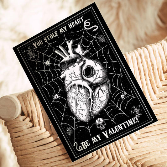 A gothic-style Valentine’s Day postcard with a black background featuring a detailed anatomical heart entangled in a spider web, surrounded by small skulls, stars, and eerie elements. The text reads 'You Stole My Heart' at the top and 'Be My Valentine?' at the bottom in a vintage-style font.