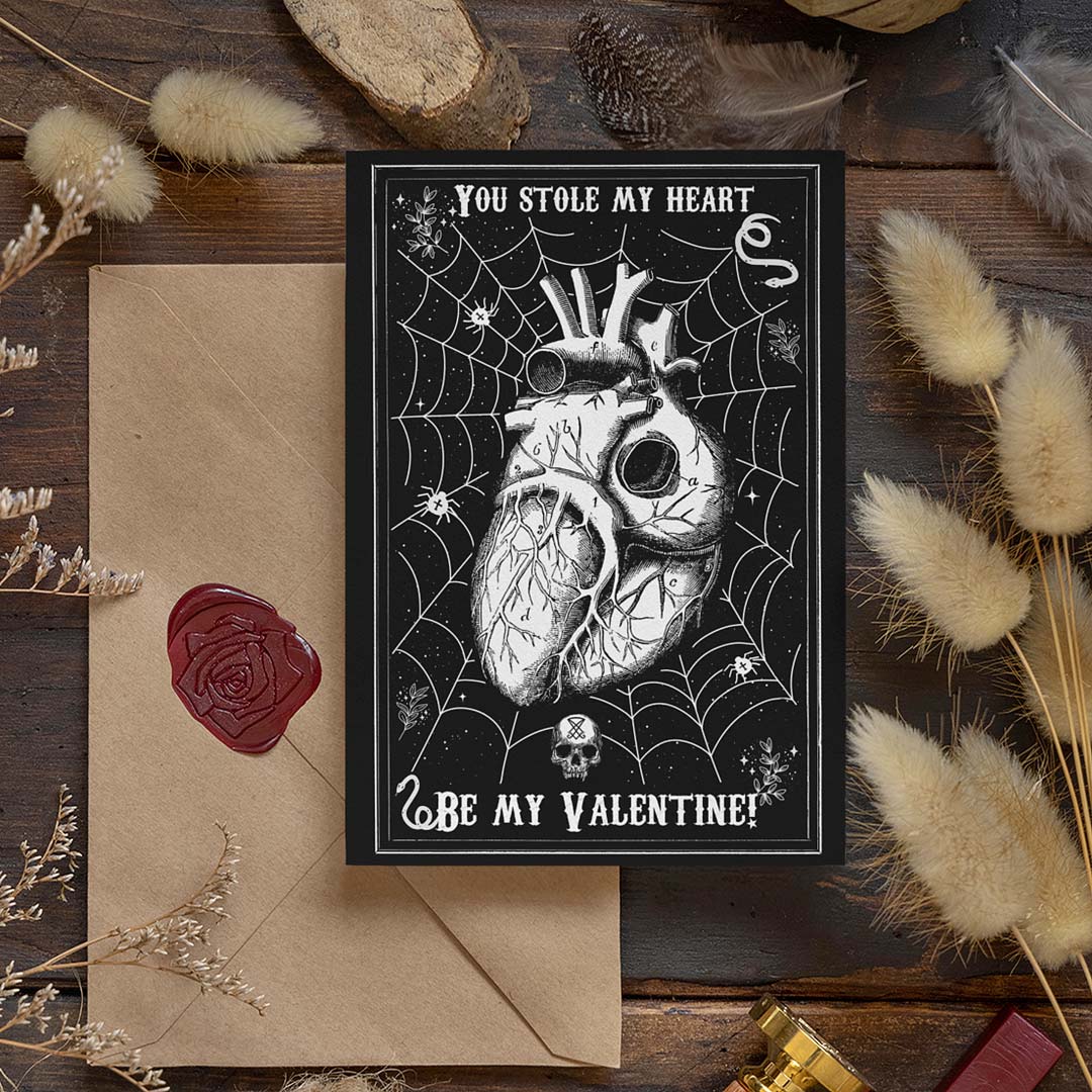 A gothic-style Valentine’s Day postcard with a black background featuring a detailed anatomical heart entangled in a spider web, surrounded by small skulls, stars, and eerie elements. The text reads 'You Stole My Heart' at the top and 'Be My Valentine?' at the bottom in a vintage-style font. The postcard is placed on a rustic wooden surface, accompanied by a kraft envelope sealed with a red wax stamp featuring a rose design. Dried flowers and soft lighting add a warm, mystical atmosphere to the scene.