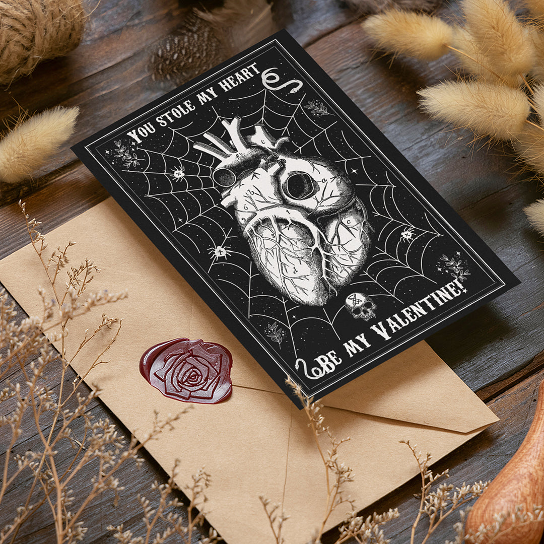 A gothic-style Valentine’s Day postcard with a black background featuring a detailed anatomical heart entangled in a spider web, surrounded by small skulls, stars, and eerie elements. The text reads 'You Stole My Heart' at the top and 'Be My Valentine?' at the bottom in a vintage-style font. The postcard is placed on a rustic wooden surface, accompanied by a kraft envelope sealed with a red wax stamp featuring a rose design. Dried flowers and soft lighting add a warm, mystical atmosphere to the scene.