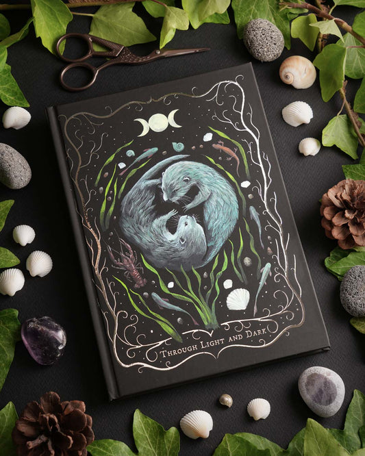 Hand-painted otter journal by Carasusan Art & Paper with silver foil details on a black A5 hardcover notebook. Nature-inspired notebook featuring moon phases and aquatic plants.
