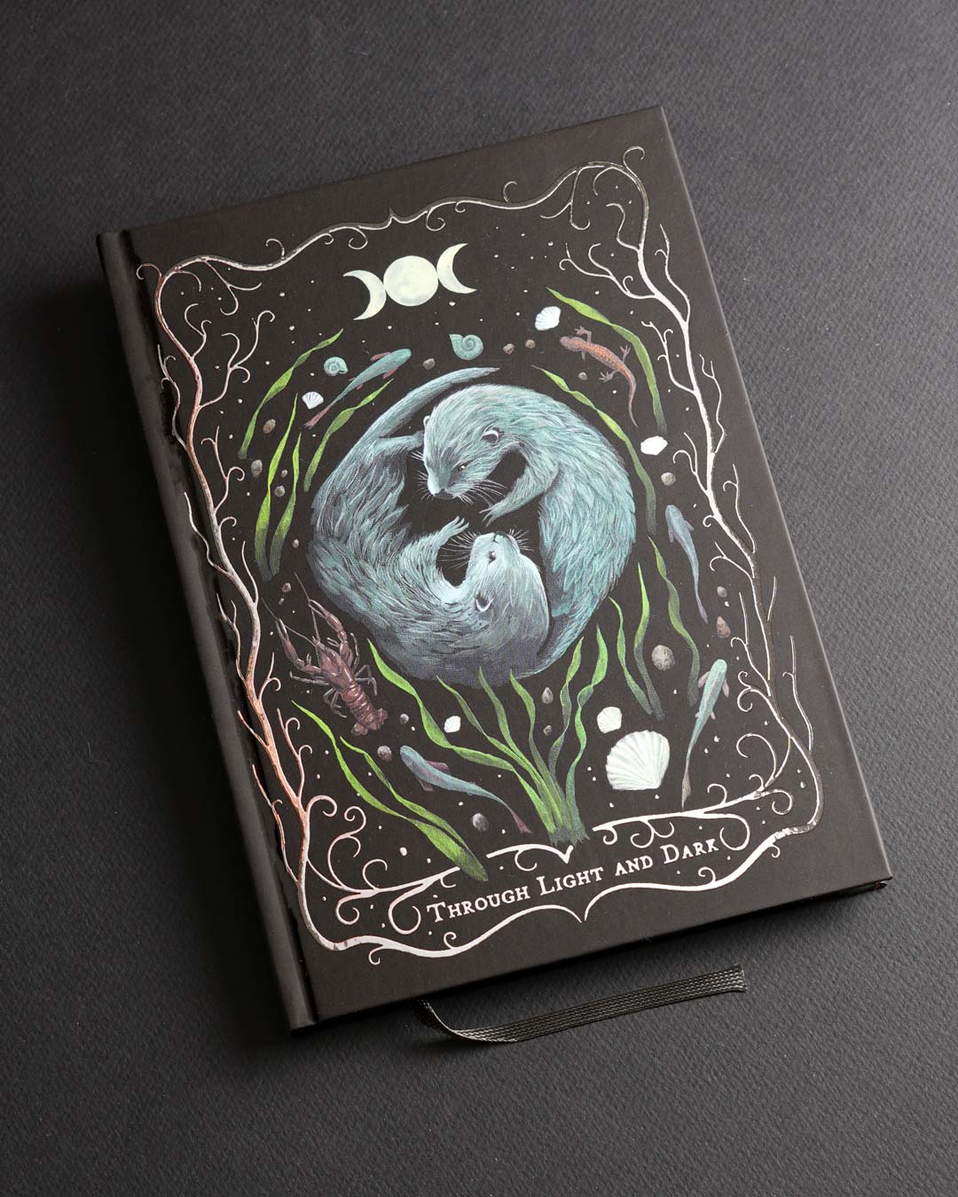 A5 hardcover notebook featuring a hand-painted illustration of two otters surrounded by water plants, shells and moon phases on a black soft-touch cover with delicate silver foil details.