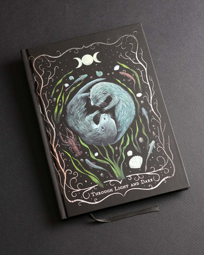 A5 hardcover notebook featuring a hand-painted illustration of two otters surrounded by water plants, shells and moon phases on a black soft-touch cover with delicate silver foil details.