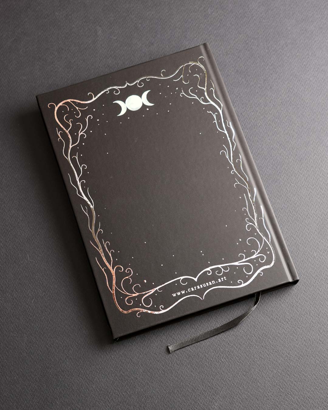 Back cover of the A5 hardcover notebook with a black soft-touch finish, elegant silver foil border featuring woodland-inspired branches, moon symbols and stars, plus a black ribbon bookmark.