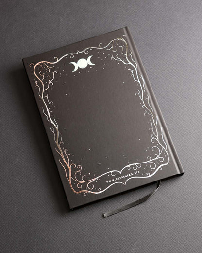 Back cover of the A5 hardcover notebook with a black soft-touch finish, elegant silver foil border featuring woodland-inspired branches, moon symbols and stars, plus a black ribbon bookmark.