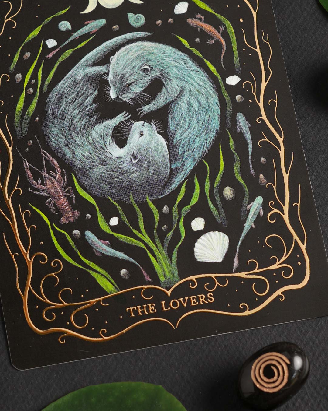 Close-up of the hand-painted "The Lovers" Tarot postcard showing the two otters and elegant gold foil details.