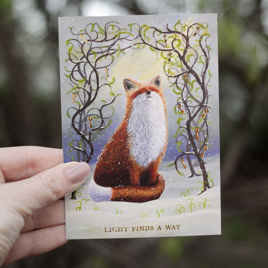 Hand holding an A6 fox postcard outdoors against a softly blurred woodland background. The artwork shows a red fox sitting in the snow beneath twisting branches with fresh spring leaves and gold foil keys. Gold foil lettering at the bottom reads “Light Finds A Way”.