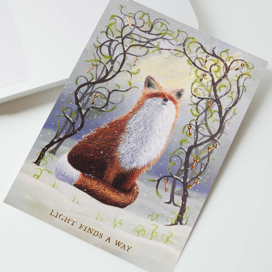 Close-up of an A6 fox postcard featuring an original oil painting of a red fox in the snow surrounded by winding branches, fresh green spring shoots and gold foil keys. The gold foil inscription “Light Finds A Way” is visible at the bottom of the artwork. Printed on premium postcard cardstock.