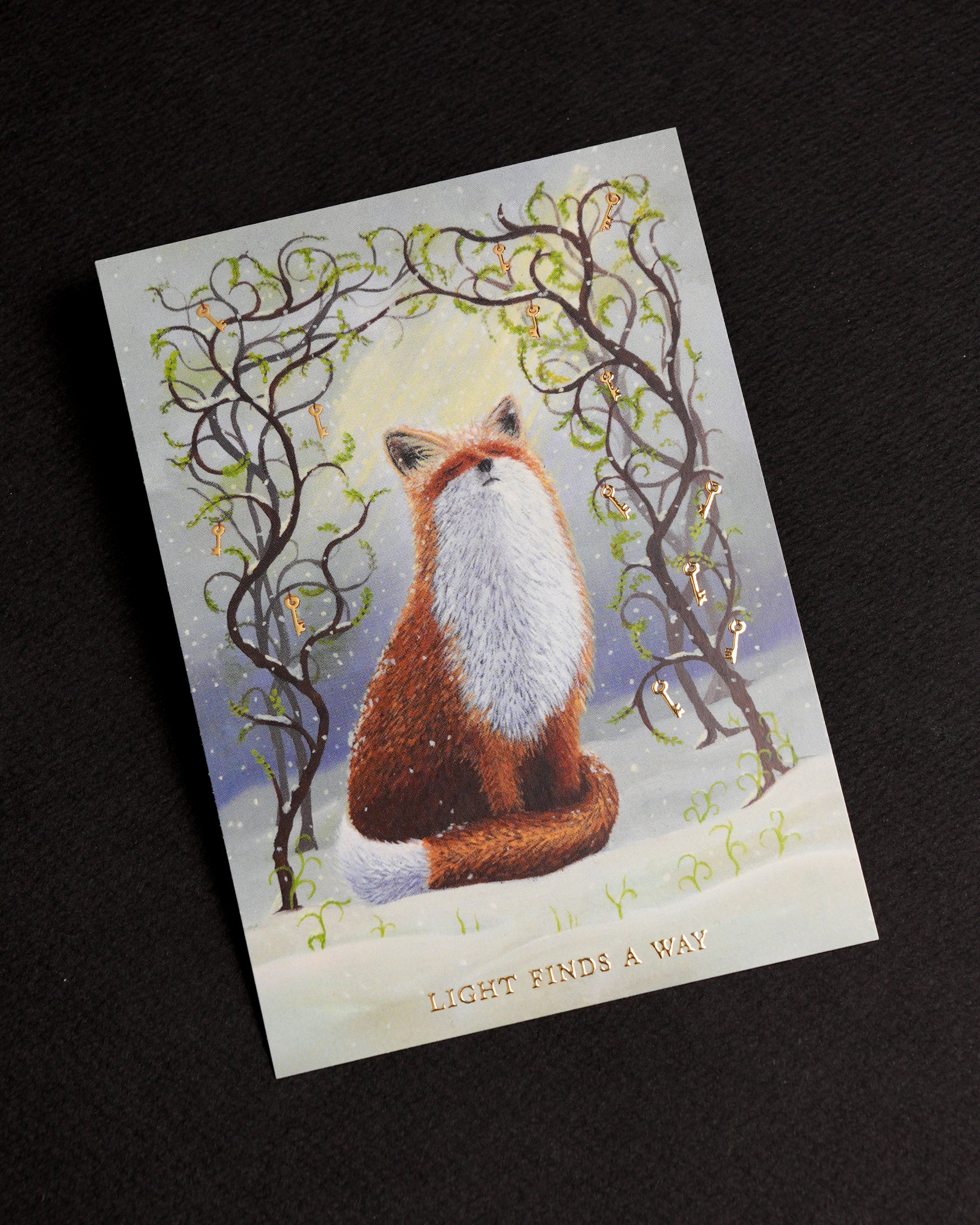 Witchy fox postcard with gold foil details displayed on a black background. The artwork features a red fox sitting in the snow beneath twisting branches adorned with golden keys and fresh green spring shoots. Gold foil lettering reads “Light Finds A Way”.