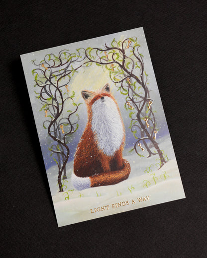 Witchy fox postcard with gold foil details displayed on a black background. The artwork features a red fox sitting in the snow beneath twisting branches adorned with golden keys and fresh green spring shoots. Gold foil lettering reads “Light Finds A Way”.