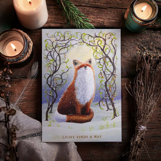Witchy fox postcard with gold foil details displayed on a rustic wooden table surrounded by candles, dried herbs and woodland treasures. The artwork features a red fox sitting in the snow beneath twisting branches adorned with golden keys and fresh green spring shoots. Gold foil lettering reads “Light Finds A Way”.