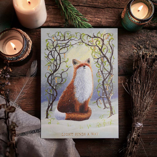 Witchy fox postcard with gold foil details displayed on a rustic wooden table surrounded by candles, dried herbs and woodland treasures. The artwork features a red fox sitting in the snow beneath twisting branches adorned with golden keys and fresh green spring shoots. Gold foil lettering reads “Light Finds A Way”.