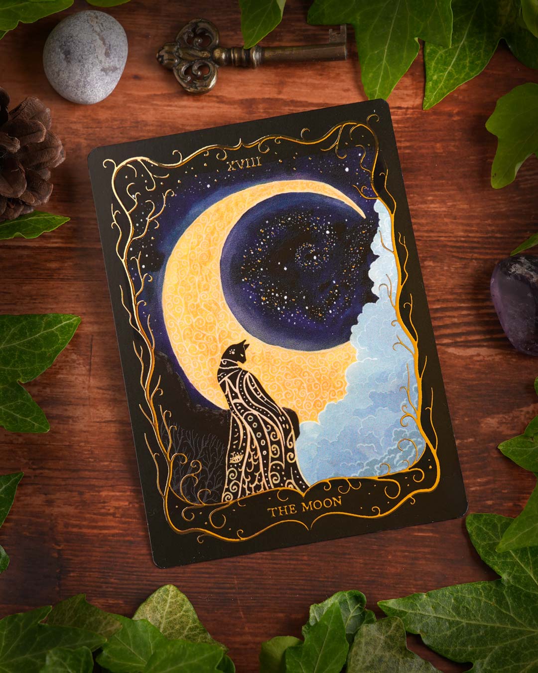 Hand-painted Tarot postcard "The Moon" with elegant gold foil details, displayed on a rustic wooden table with ivy, an antique key and natural decorations.