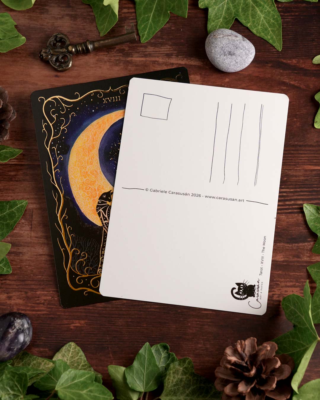 Front and back of the "The Moon" Tarot postcard featuring gold foil details and a classic postcard back, styled on a wooden table with ivy.