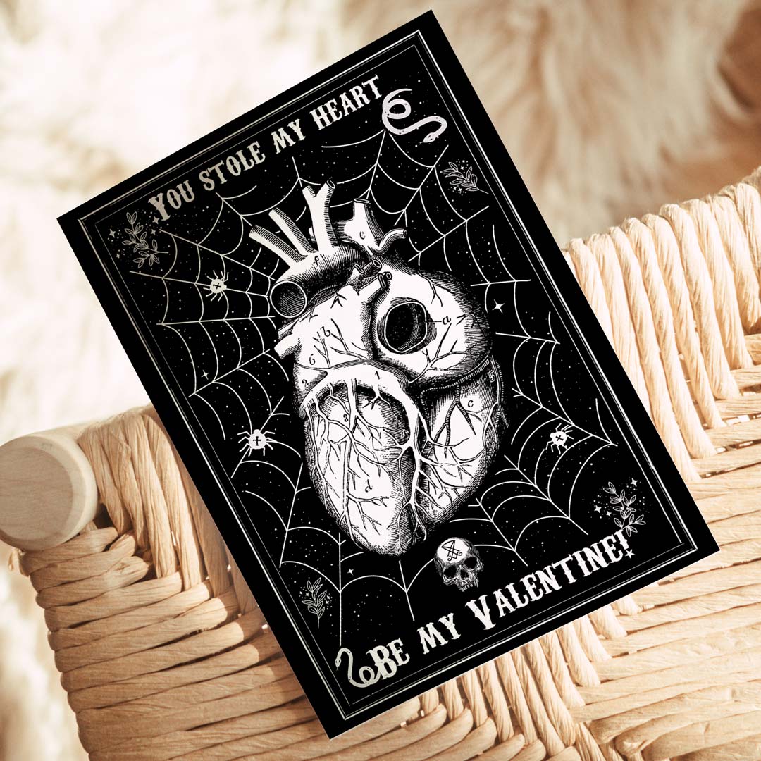 A gothic-style Valentine’s Day postcard with a black background featuring a detailed anatomical heart entangled in a spider web, surrounded by small skulls, stars, and eerie elements. The text reads 'You Stole My Heart' at the top and 'Be My Valentine?' at the bottom in a vintage-style font.